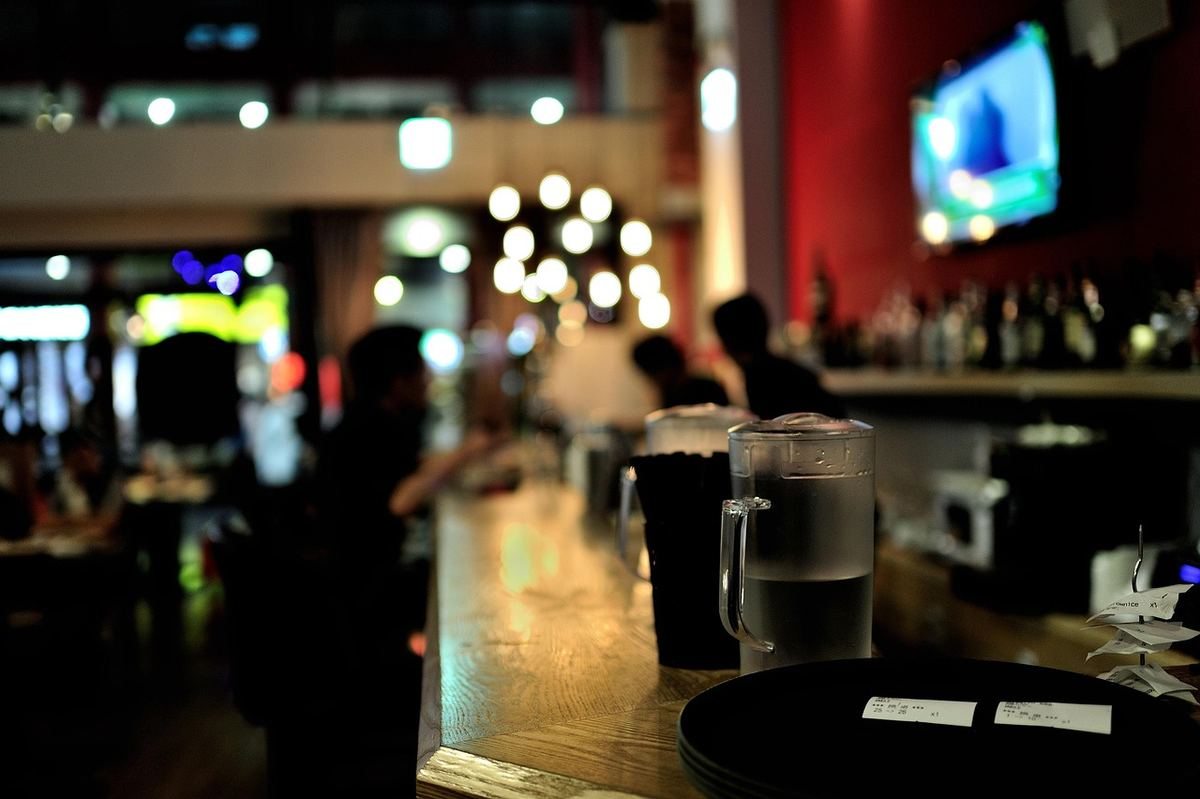 Comptoir de bar éclairé avec verres, clients flous en arrière-plan, écrans lumineux visibles